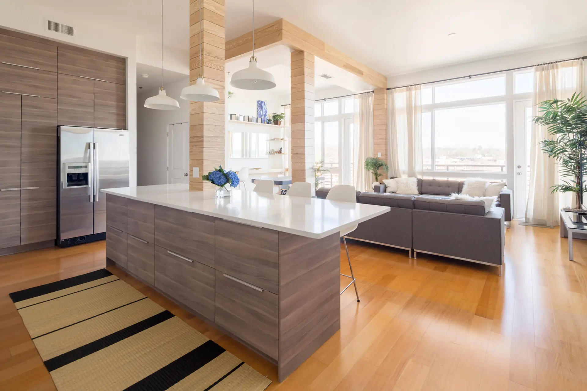 Modern kitchen and living area with wood floors, large island, gray cabinets, stainless steel fridge, and a gray sectional so