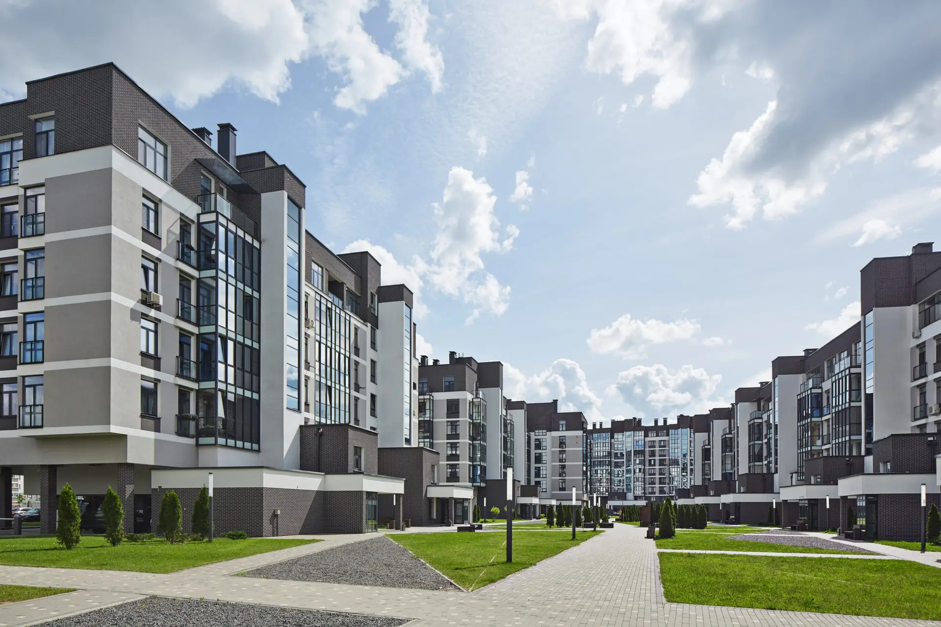 Modern apartment buildings with large windows, surrounded by green lawns and a paved walkway under a cloudy sky.