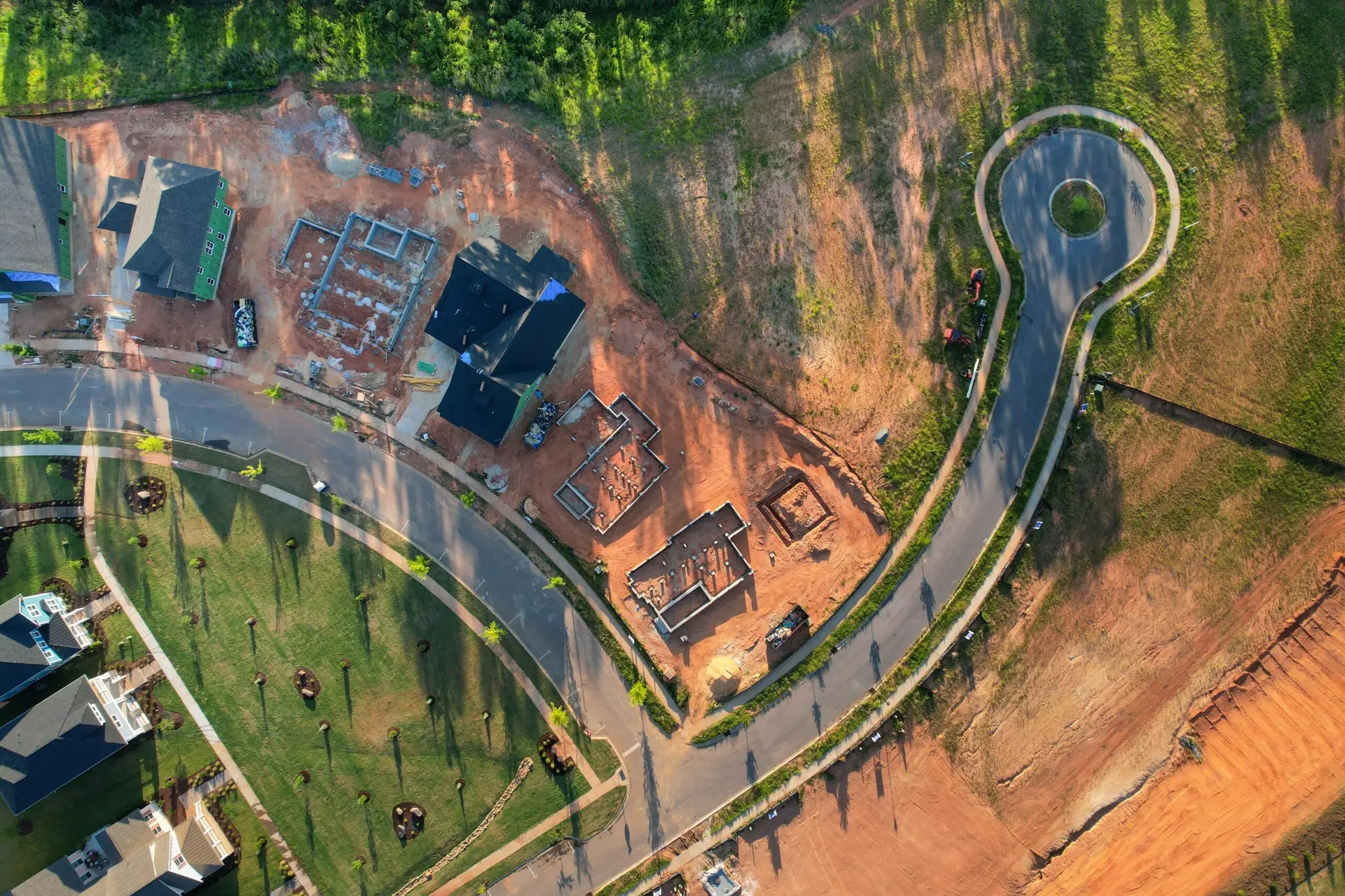 Aerial view of a residential construction site with houses, foundations, and a circular road surrounded by greenery.