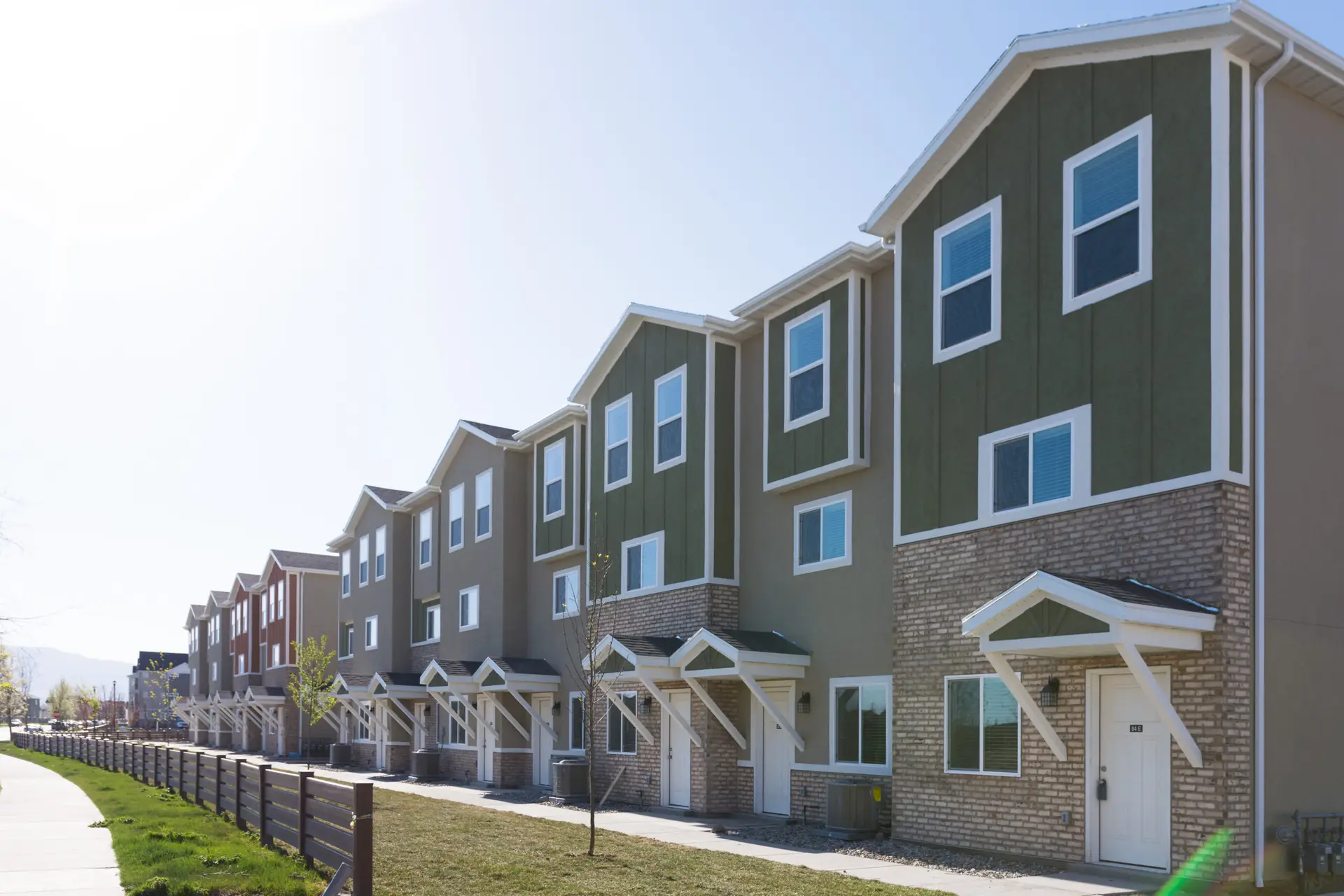 Row of modern townhouses with brick and green siding, white trim, and small front porches along a sidewalk.