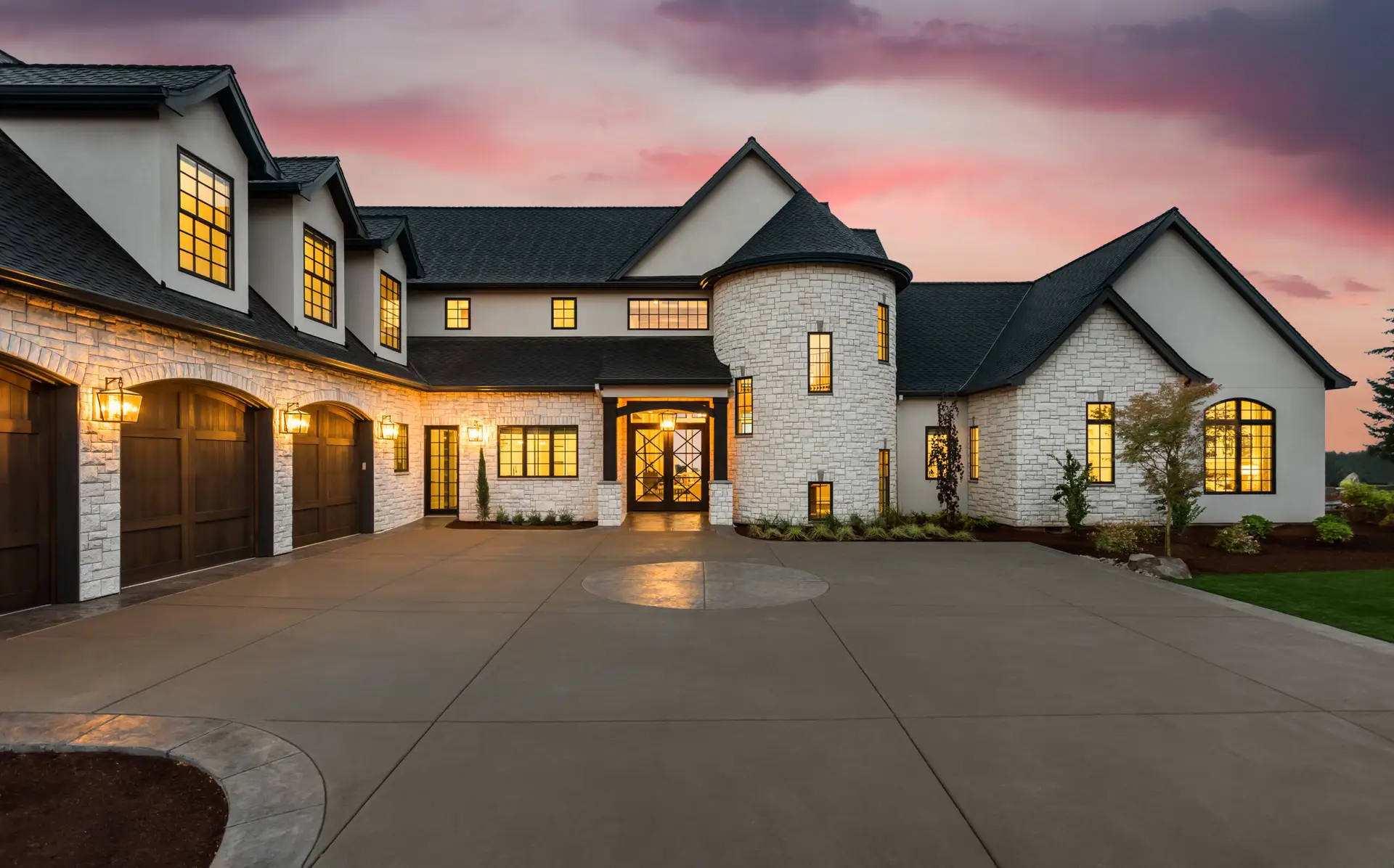 Elegant stone house with large windows, circular turret, and lit exterior at sunset.