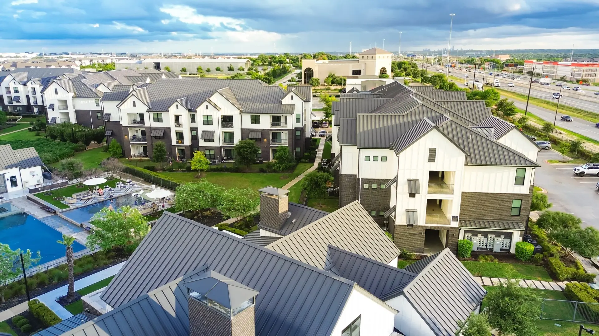 Modern apartment complex with landscaped gardens, swimming pool, and nearby highway under a cloudy sky.