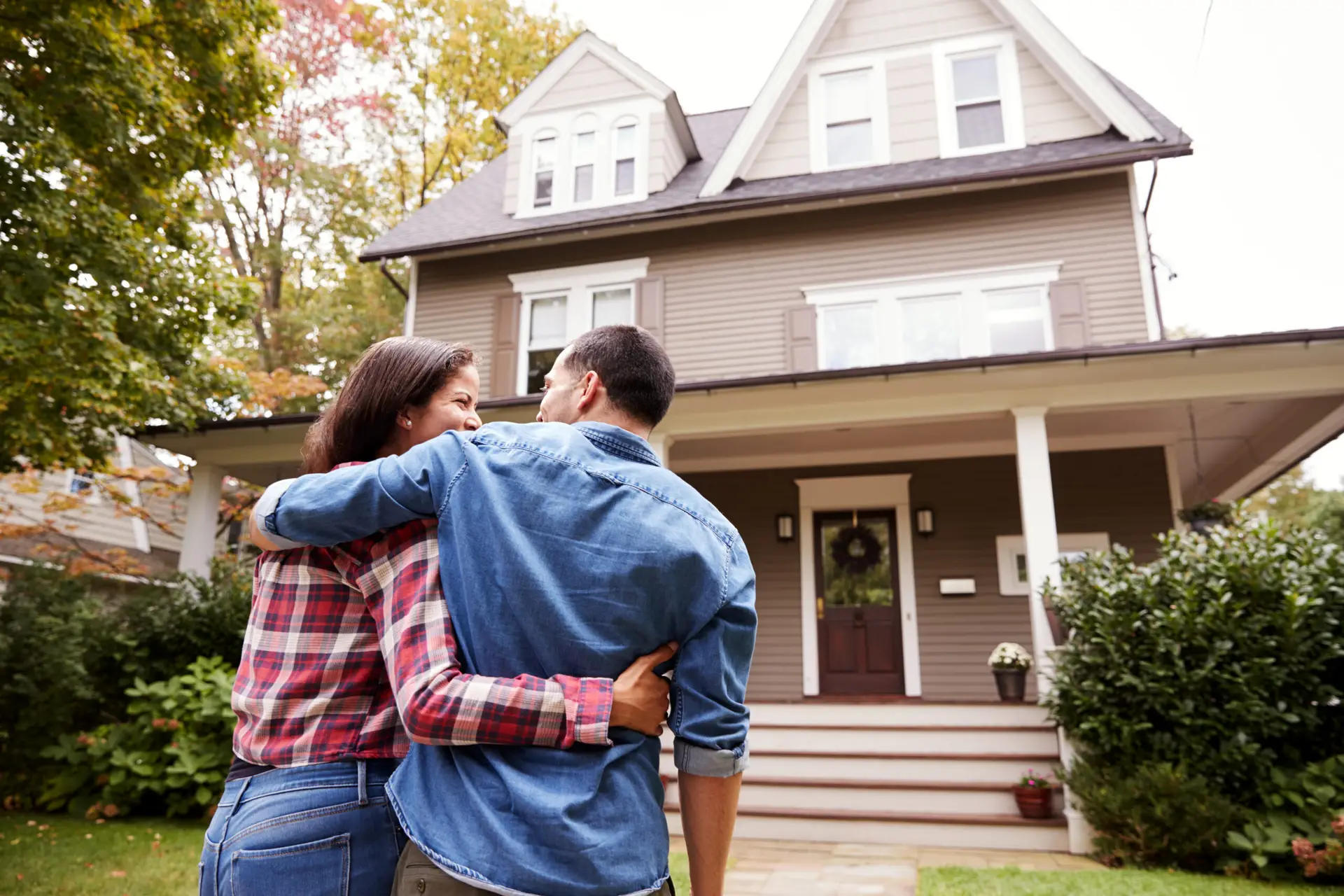 Couple embracing while looking at a two-story house with a porch and greenery.