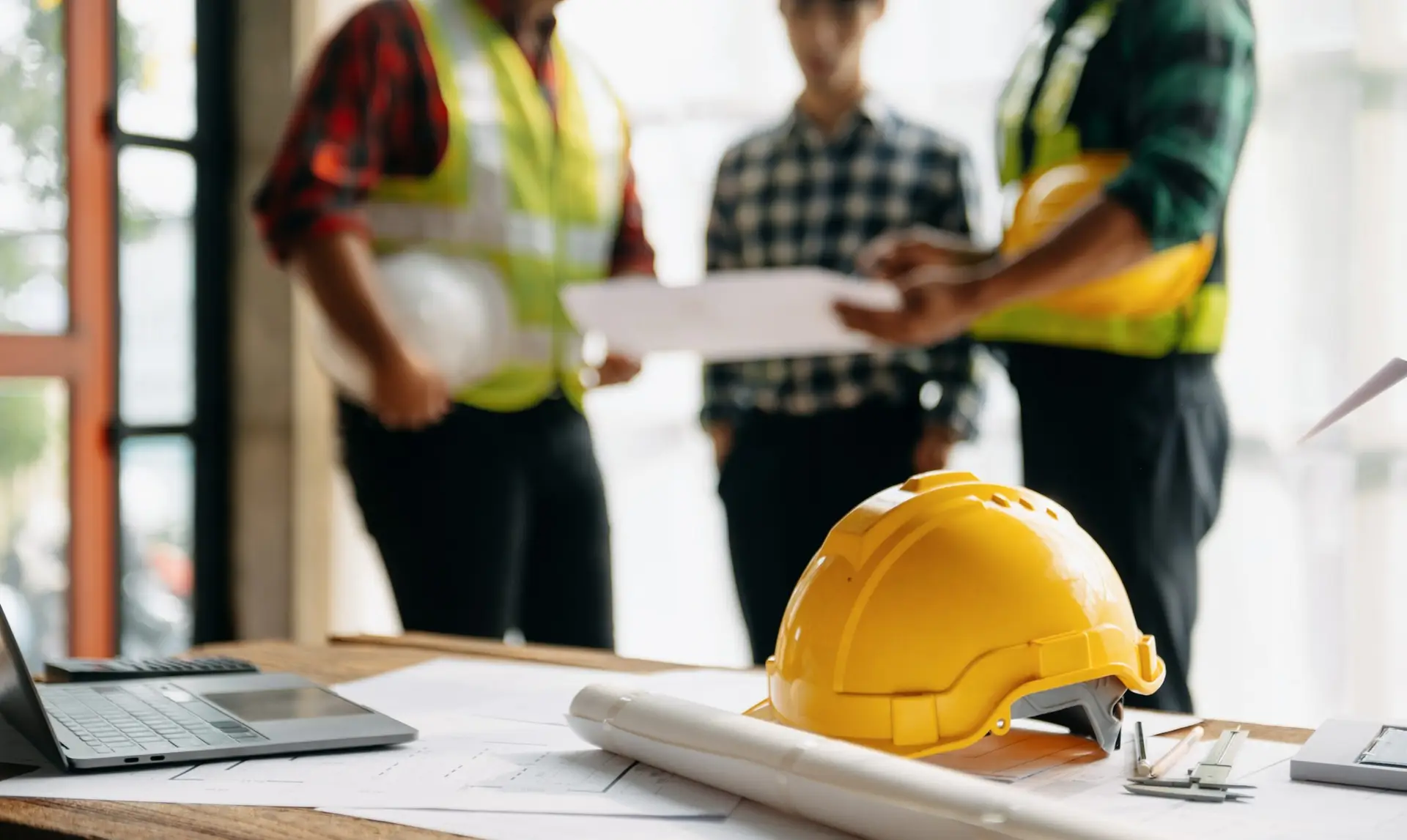 Yellow hard hat on table with blueprints, laptop, and three construction workers discussing plans in the background.